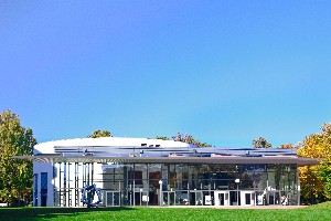 Modernes, glasumhülltes Gebäude vor blauem Himmel und herbstlichen Bäumen im Park.